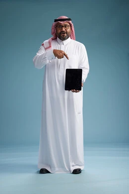 A Saudi man in traditional attire standing using a tablet, using the tablet, blue background, full body.