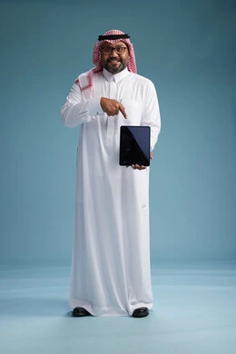 A Saudi man in traditional attire standing using a tablet, using the tablet, blue background, full body.