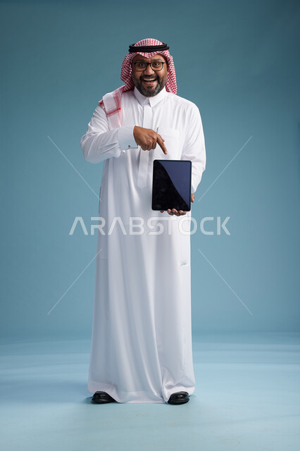 رجل سعودي بالزي التقليدي واقف يستخدم جهاز لوحي, استخدام جهاز التابليت, خلفيه زرقاء, عرض شاشه سوداء فارغه, كامل الجسم