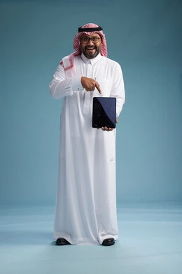 A Saudi man in traditional attire standing using a tablet, using the tablet, blue background, full body.
