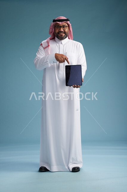 A Saudi man in traditional attire standing using a tablet, using the tablet, blue background, full body.