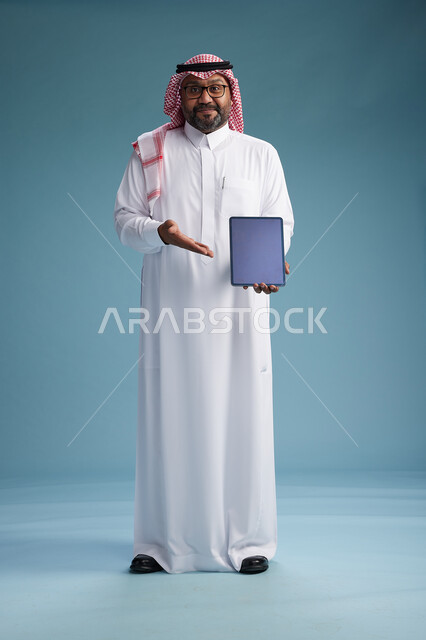 A Saudi man in traditional attire standing using a tablet, using the tablet, blue background, full body.