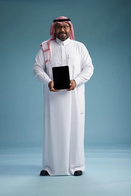 A Saudi man in traditional attire standing using a tablet, using the tablet, blue background, full body.