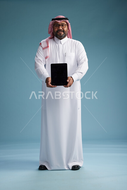 رجل سعودي بالزي التقليدي واقف يستخدم جهاز لوحي, استخدام جهاز التابليت, خلفيه زرقاء, عرض شاشه سوداء فارغه, كامل الجسم