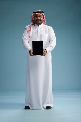 رجل سعودي بالزي التقليدي واقف يستخدم جهاز لوحي, استخدام جهاز التابليت, خلفيه زرقاء, عرض شاشه سوداء فارغه, كامل الجسم