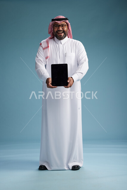 A Saudi man in traditional attire standing using a tablet, using the tablet, blue background, full body.