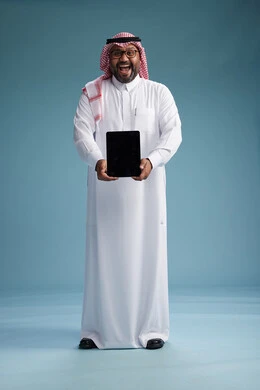 A Saudi man in traditional attire standing using a tablet, using the tablet, blue background, full body.