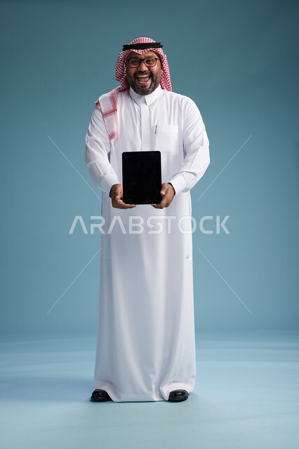 A Saudi man in traditional attire standing using a tablet, using the tablet, blue background, full body.