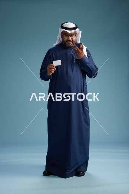 بورتريه لرجل سعودي يرتدي ثوب كحلي يقوم بعملية دفع عبر الهاتف المحمول, مفهوم الشراء الاونلاين, كامل الجسم, خلفيه زرقاء