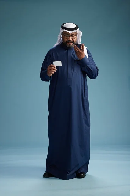 A portrait of a Saudi man wearing a navy thobe making a payment via mobile phone, the concept of online shopping, full body, blue background.