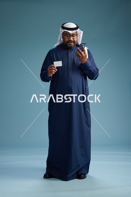 بورتريه لرجل سعودي يرتدي ثوب كحلي يقوم بعملية دفع عبر الهاتف المحمول, مفهوم الشراء الاونلاين, كامل الجسم, خلفيه زرقاء