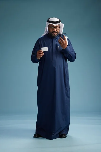 بورتريه لرجل سعودي يرتدي ثوب كحلي يقوم بعملية دفع عبر الهاتف المحمول, مفهوم الشراء الاونلاين, كامل الجسم, خلفيه زرقاء