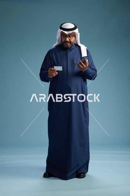 بورتريه لرجل سعودي يرتدي ثوب كحلي يقوم بعملية دفع عبر الهاتف المحمول, مفهوم الشراء الاونلاين, كامل الجسم, خلفيه زرقاء