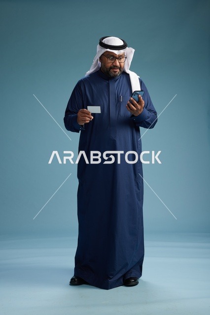 A portrait of a Saudi man wearing a navy thobe making a payment via mobile phone, the concept of online shopping, full body, blue background.