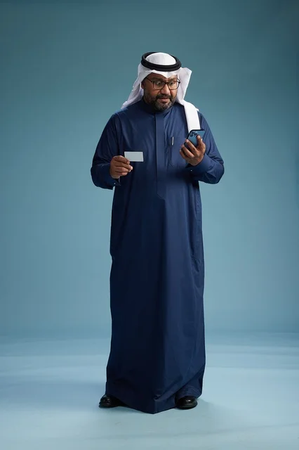 بورتريه لرجل سعودي يرتدي ثوب كحلي يقوم بعملية دفع عبر الهاتف المحمول, مفهوم الشراء الاونلاين, كامل الجسم, خلفيه زرقاء