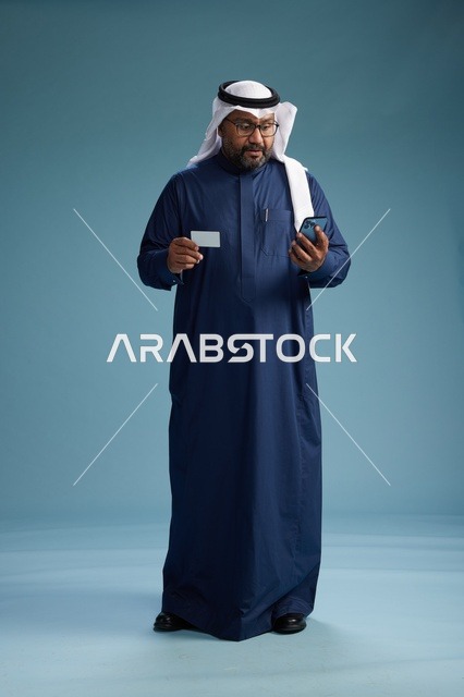 بورتريه لرجل سعودي يرتدي ثوب كحلي يقوم بعملية دفع عبر الهاتف المحمول, مفهوم الشراء الاونلاين, كامل الجسم, خلفيه زرقاء