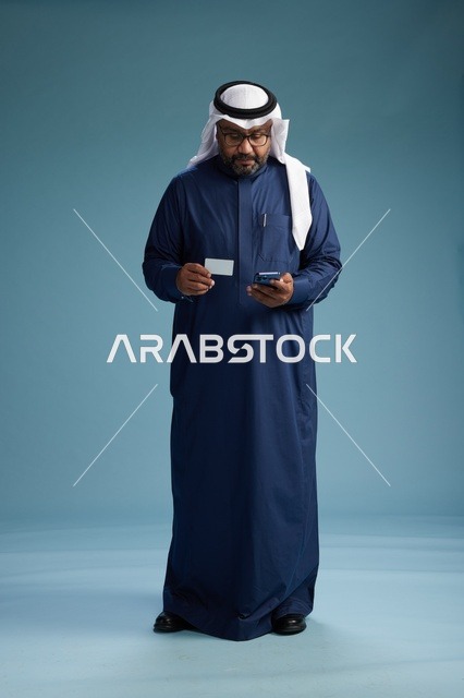 بورتريه لرجل سعودي يرتدي ثوب كحلي يقوم بعملية دفع عبر الهاتف المحمول, مفهوم الشراء الاونلاين, كامل الجسم, خلفيه زرقاء