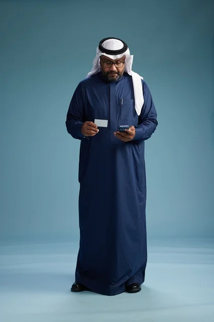 بورتريه لرجل سعودي يرتدي ثوب كحلي يقوم بعملية دفع عبر الهاتف المحمول, مفهوم الشراء الاونلاين, كامل الجسم, خلفيه زرقاء