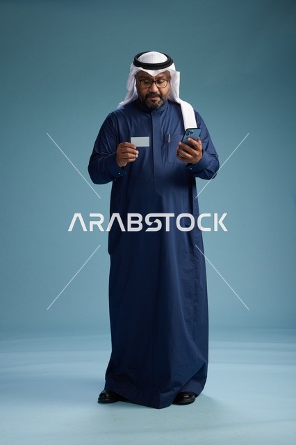 بورتريه لرجل سعودي يرتدي ثوب كحلي يقوم بعملية دفع عبر الهاتف المحمول, مفهوم الشراء الاونلاين, كامل الجسم, خلفيه زرقاء