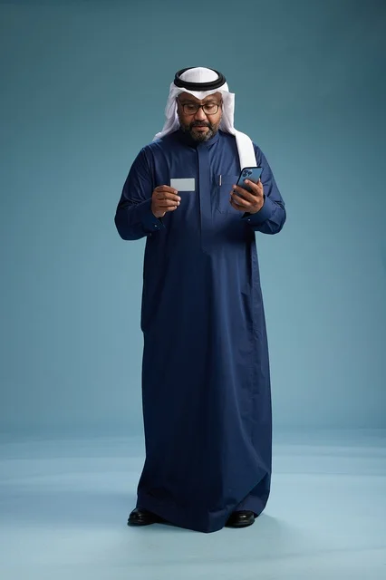 بورتريه لرجل سعودي يرتدي ثوب كحلي يقوم بعملية دفع عبر الهاتف المحمول, مفهوم الشراء الاونلاين, كامل الجسم, خلفيه زرقاء