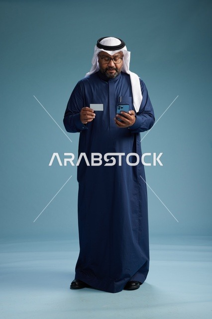 بورتريه لرجل سعودي يرتدي ثوب كحلي يقوم بعملية دفع عبر الهاتف المحمول, مفهوم الشراء الاونلاين, كامل الجسم, خلفيه زرقاء