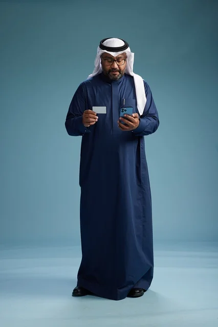 بورتريه لرجل سعودي يرتدي ثوب كحلي يقوم بعملية دفع عبر الهاتف المحمول, مفهوم الشراء الاونلاين, كامل الجسم, خلفيه زرقاء