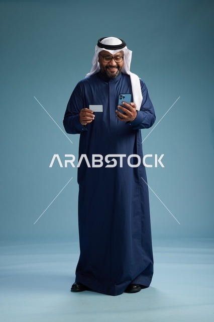 A portrait of a Saudi man wearing a navy thobe making a payment via mobile phone, the concept of online shopping, full body, blue background.