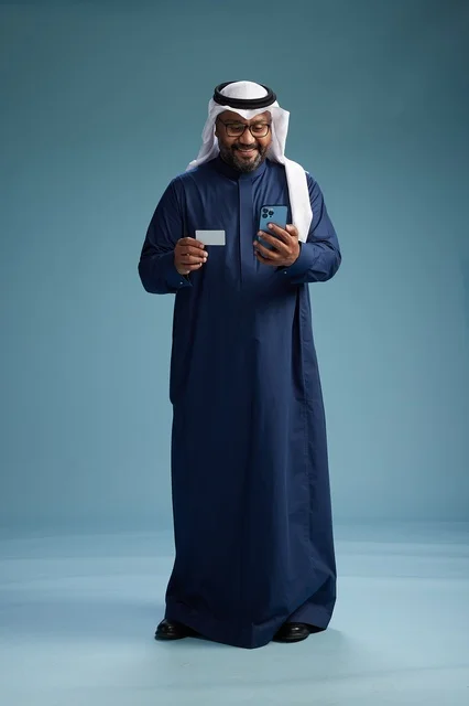 بورتريه لرجل سعودي يرتدي ثوب كحلي يقوم بعملية دفع عبر الهاتف المحمول, مفهوم الشراء الاونلاين, كامل الجسم, خلفيه زرقاء