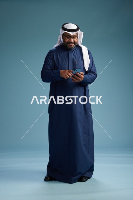 بورتريه لرجل سعودي يرتدي ثوب كحلي يقوم بعملية دفع عبر الهاتف المحمول, مفهوم الشراء الاونلاين, كامل الجسم, خلفيه زرقاء
