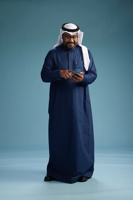 بورتريه لرجل سعودي يرتدي ثوب كحلي يقوم بعملية دفع عبر الهاتف المحمول, مفهوم الشراء الاونلاين, كامل الجسم, خلفيه زرقاء