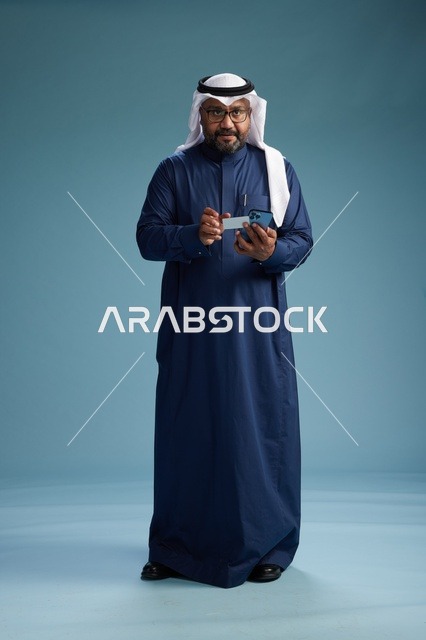 A portrait of a Saudi man wearing a navy thobe making a payment via mobile phone, the concept of online shopping, full body, blue background.