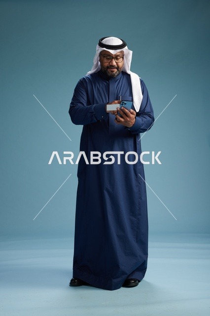 بورتريه لرجل سعودي يرتدي ثوب كحلي يقوم بعملية دفع عبر الهاتف المحمول, مفهوم الشراء الاونلاين, كامل الجسم, خلفيه زرقاء