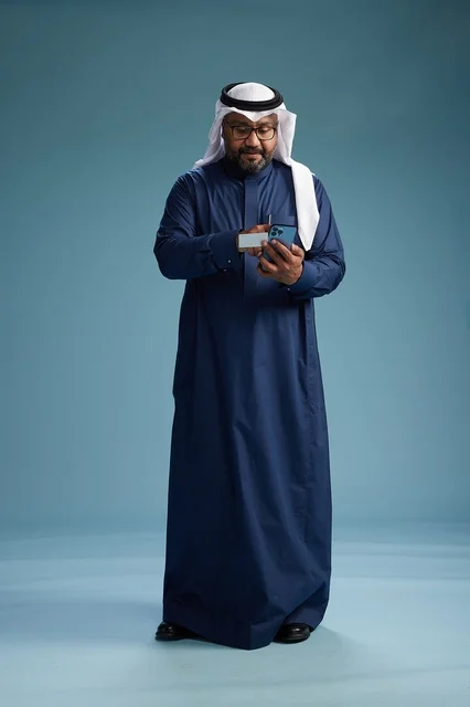 بورتريه لرجل سعودي يرتدي ثوب كحلي يقوم بعملية دفع عبر الهاتف المحمول, مفهوم الشراء الاونلاين, كامل الجسم, خلفيه زرقاء
