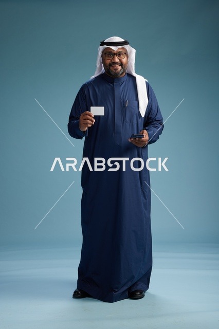 بورتريه لرجل سعودي يرتدي ثوب كحلي يقوم بعملية دفع عبر الهاتف المحمول, مفهوم الشراء الاونلاين, كامل الجسم, خلفيه زرقاء