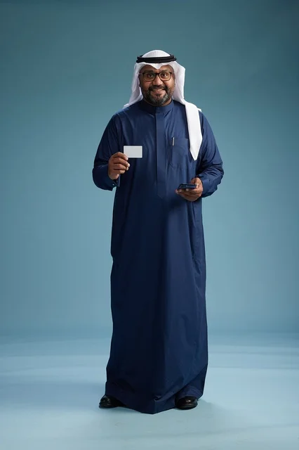 A portrait of a Saudi man wearing a navy thobe making a payment via mobile phone, the concept of online shopping, full body, blue background.