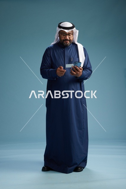 بورتريه لرجل سعودي يرتدي ثوب كحلي يقوم بعملية دفع عبر الهاتف المحمول, مفهوم الشراء الاونلاين, كامل الجسم, خلفيه زرقاء