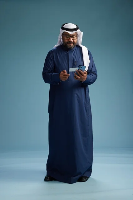 بورتريه لرجل سعودي يرتدي ثوب كحلي يقوم بعملية دفع عبر الهاتف المحمول, مفهوم الشراء الاونلاين, كامل الجسم, خلفيه زرقاء