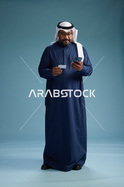 A portrait of a Saudi man wearing a navy thobe making a payment via mobile phone, the concept of online shopping, full body, blue background.