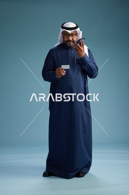بورتريه لرجل سعودي يرتدي ثوب كحلي يقوم بعملية دفع عبر الهاتف المحمول, مفهوم الشراء الاونلاين, كامل الجسم, خلفيه زرقاء