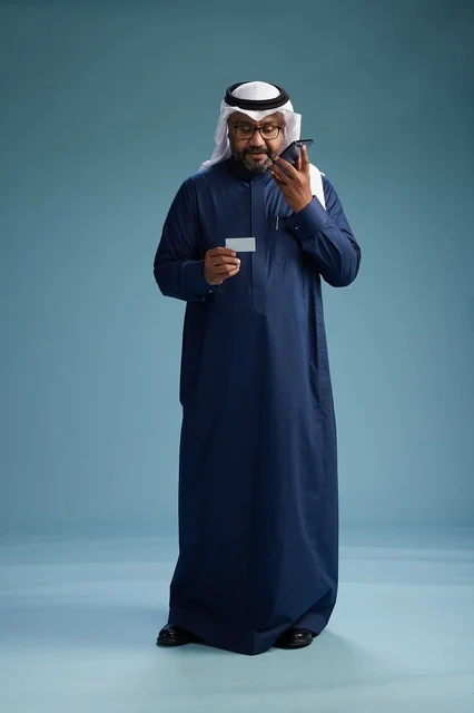 بورتريه لرجل سعودي يرتدي ثوب كحلي يقوم بعملية دفع عبر الهاتف المحمول, مفهوم الشراء الاونلاين, كامل الجسم, خلفيه زرقاء