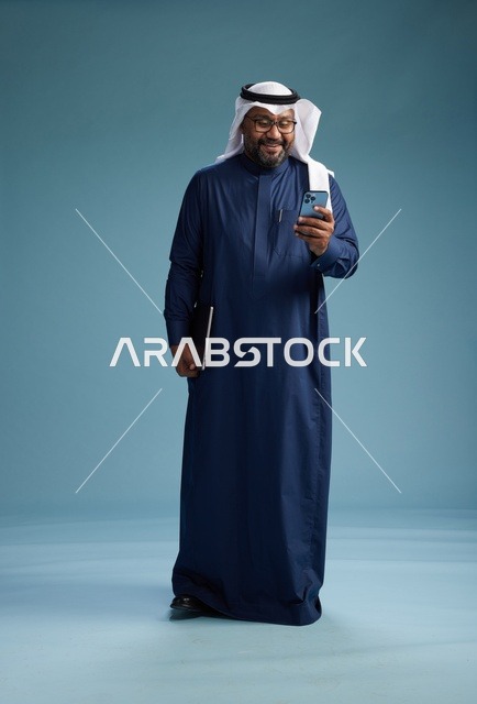 بورتريه لرجل سعودي مبتسم  يرتدي ثوب كحلي وغتره بيضاء يحمل لابتوب وهاتف ذكي, استخدام التقنيه الحديثه, كامل الجسم, خلفيه زرقاء