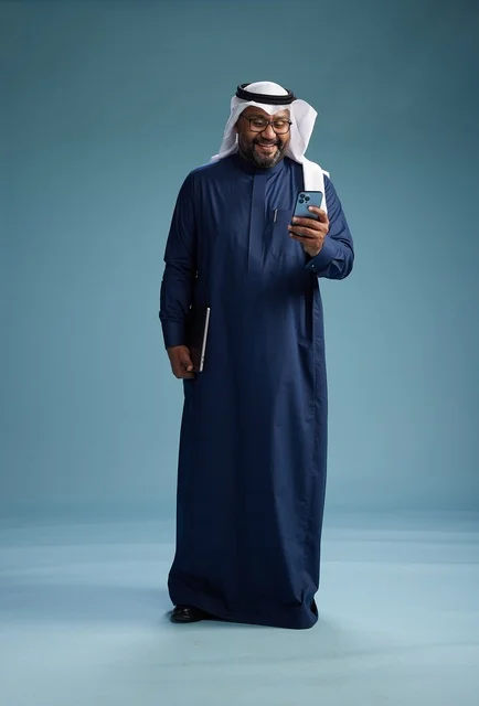 A portrait of a smiling Saudi man wearing a navy blue thobe and a white ghutrah, holding a laptop and a smartphone, using modern technology, full body, with a blue background.