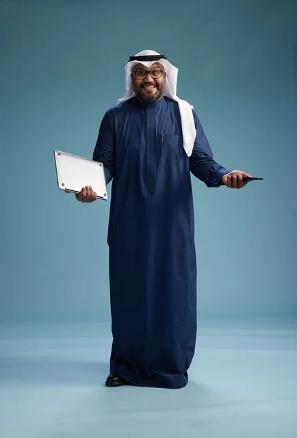A portrait of a smiling Saudi man wearing a navy blue thobe and a white ghutrah, holding a laptop and a smartphone, using modern technology, full body, with a blue background.