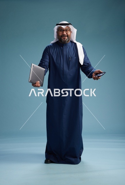 بورتريه لرجل سعودي مبتسم  يرتدي ثوب كحلي وغتره بيضاء يحمل لابتوب وهاتف ذكي, استخدام التقنيه الحديثه, كامل الجسم, خلفيه زرقاء
