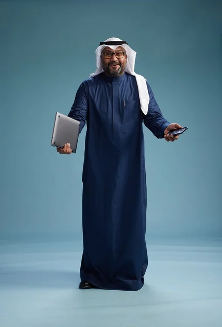 A portrait of a smiling Saudi man wearing a navy blue thobe and a white ghutrah, holding a laptop and a smartphone, using modern technology, full body, with a blue background.