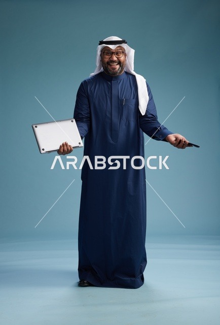 بورتريه لرجل سعودي مبتسم  يرتدي ثوب كحلي وغتره بيضاء يحمل لابتوب وهاتف ذكي, استخدام التقنيه الحديثه, كامل الجسم, خلفيه زرقاء