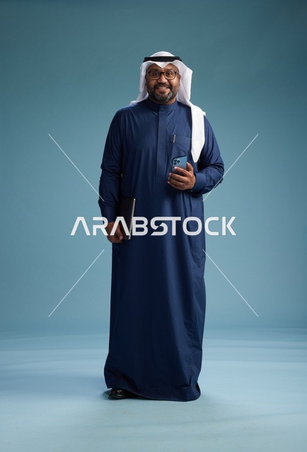 بورتريه لرجل سعودي مبتسم  يرتدي ثوب كحلي وغتره بيضاء يحمل لابتوب وهاتف ذكي, استخدام التقنيه الحديثه, كامل الجسم, خلفيه زرقاء