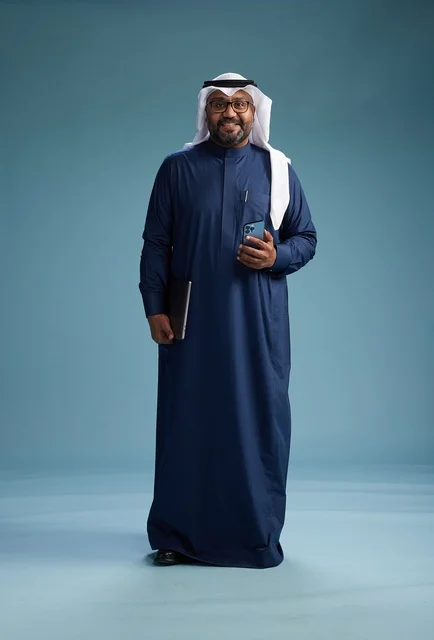 A portrait of a smiling Saudi man wearing a navy blue thobe and a white ghutrah, holding a laptop and a smartphone, using modern technology, full body, with a blue background.