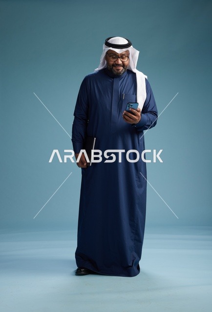 بورتريه لرجل سعودي مبتسم  يرتدي ثوب كحلي وغتره بيضاء يحمل لابتوب وهاتف ذكي, استخدام التقنيه الحديثه, كامل الجسم, خلفيه زرقاء