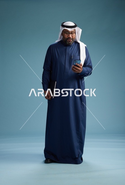 بورتريه لرجل سعودي مبتسم  يرتدي ثوب كحلي وغتره بيضاء يحمل لابتوب وهاتف ذكي, استخدام التقنيه الحديثه, كامل الجسم, خلفيه زرقاء